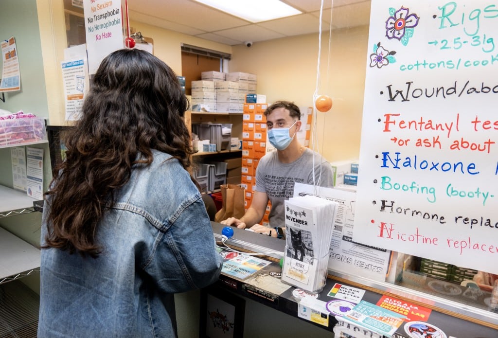 A window and counter at a syringe services program with a sign on the right that markets available services, like “rigs,” “wound” care, “fentanyl tests,” “Naloxone,” “Boofing,” “Hormone replacement,” and “Nicotine replacement.” Behind the counter is a staff person wearing a blue surgical mask and typing on a keyboard while looking at the client across the counter, who has long dark hair and a blue shirt. The counter is covered in colorful stickers.