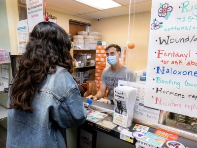 A window and counter at a syringe services program with a sign on the right that markets available services, like “rigs,” “wound” care, “fentanyl tests,” “Naloxone,” “Boofing,” “Hormone replacement,” and “Nicotine replacement.” Behind the counter is a staff person wearing a blue surgical mask and typing on a keyboard while looking at the client across the counter, who has long dark hair and a blue shirt. The counter is covered in colorful stickers.